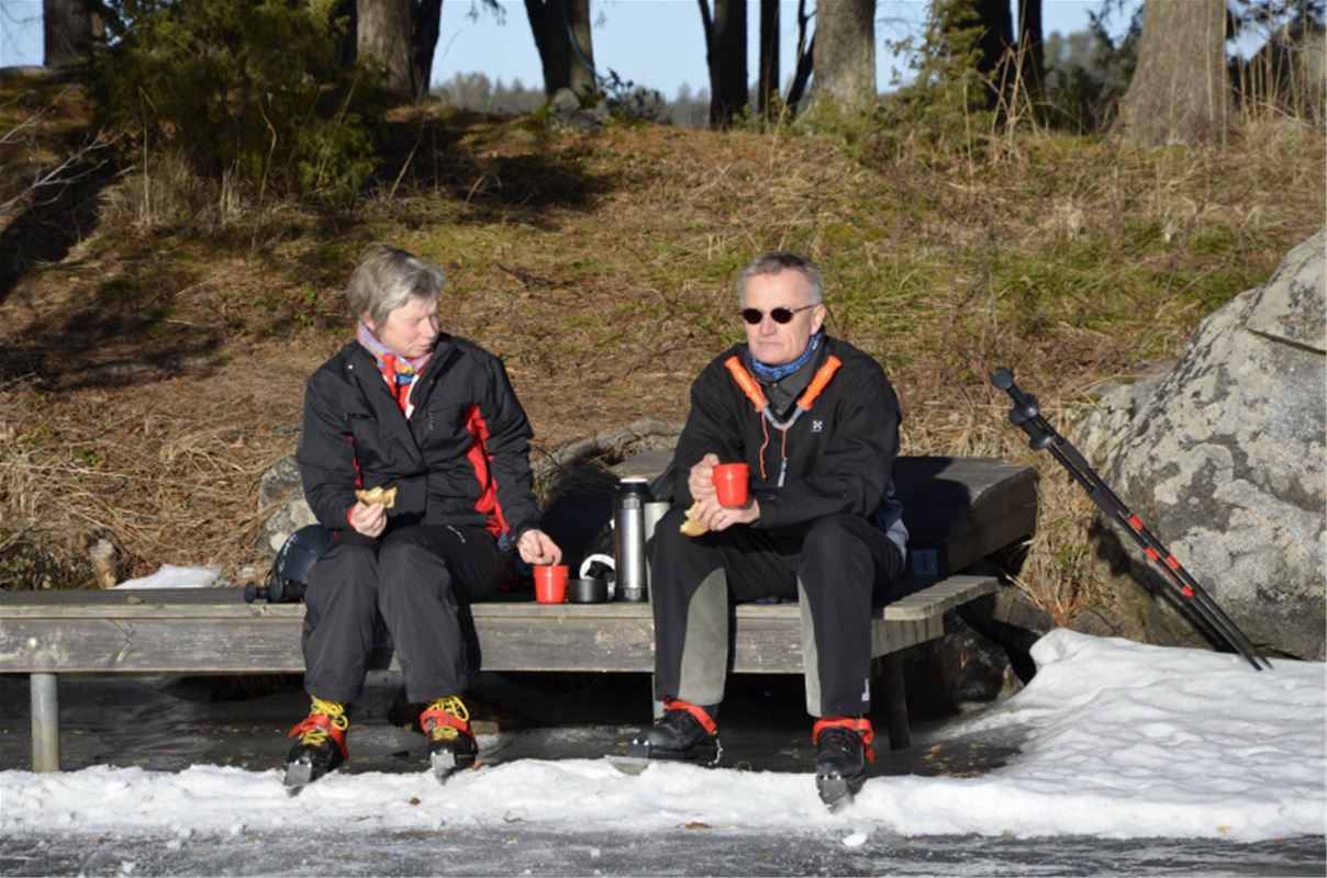 Två personer som fikar i solen och sitter på en bänk vid sjökanten som består av snö och is. 