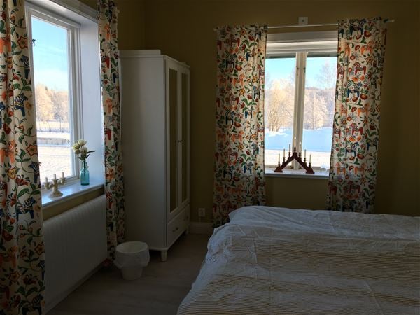 Sovrum med mönstrade gardiner med dala-motiv, vita sängkläder och vit garderob med spegeldörrar. 