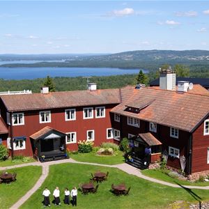 Villa Långbers från ovan en solig sommardag med sjön  Siljan i bakgrunden och en flaggstång framför den faluröda träbyggnaden.