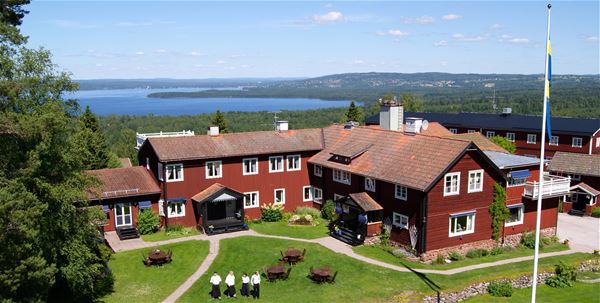 Villa Långbers från ovan en solig sommardag med sjön  Siljan i bakgrunden och en flaggstång framför den faluröda träbyggnaden. 