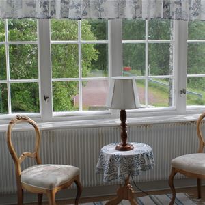 Spröjsade fönster med två stolar i trä och ett litet bord med duk och lampa framför fönstret. 