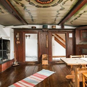 Rum med dalmålning i taket, öppen spis, matbord med stolar samt ett gammalt väggfast skåp.