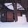 A small timbered shelter with snow on the roof and on the ground.