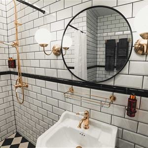 Badrum med kaklade, vita väggar och schackrutigt kaklat golv, mässingsfärgade detaljer och en svart, rund spegel. 