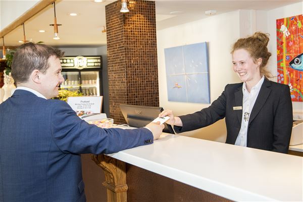 En gäst i blå kostym tar emot ett nyckelkort från en receptionist i svart kavaj.  