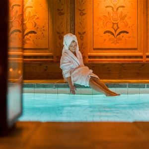 Person in a dressing-gown is sitting on the edge to the indoor pool.