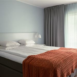 Dubbelsäng med ett orangefärgat överkast och ett fönster med gråa gardiner. 