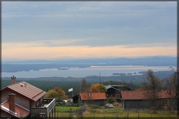 Vy över Fryksås med gråa fäbodstugor i förgrunden och Orsasjön i bakgrunden. 