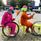 Litteraturhuset Trampolin,  © Trampolin, Två personer med färgglada overaller och kepsar leker cykel med pallar och cykelhjul.