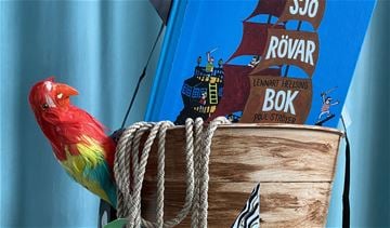 Ett foto med en röd papegoja sittandes i en mast med ett rep och en sjörövarbok.