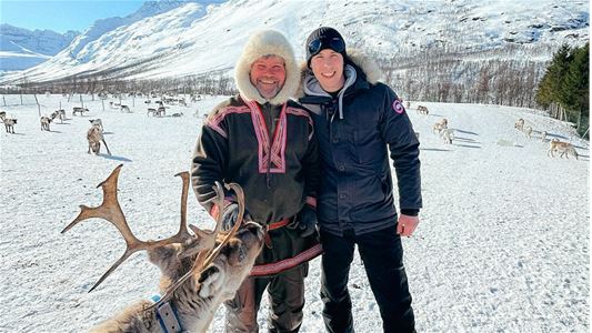 A Sami guide standing with a guest while a reindeer is looking at them