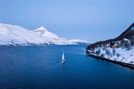 Sailboat on calm winter water surrounded by snow-covered mountains and trees, with a house by the shoreline
