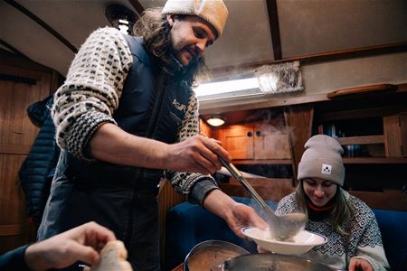 Three people in a boat cabin, one serving food from a pot