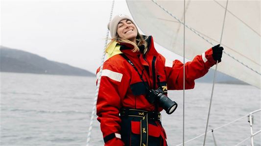 Person in sailing gear standing on a sailboat with a camera around their neck, water and mountains in the background