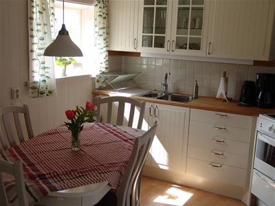 Vitt kök och ett matbord med 4 stolar och en rödrutig duk och blommor i en vas på bordet. 