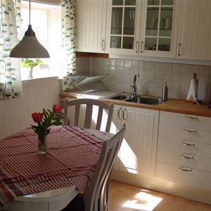Vitt kök och ett matbord med 4 stolar och en rödrutig duk och blommor i en vas på bordet. 