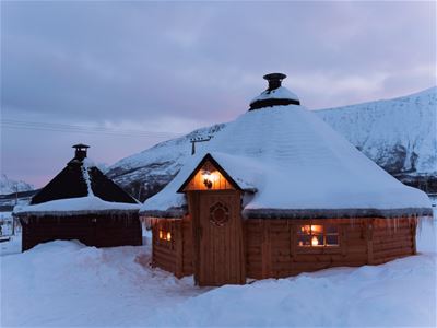 Two cabins with lights in the windows in a snowy mountain landscape at dusk.