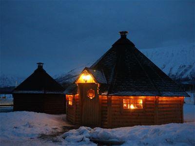 Opplyst tretakshytte i snødekt landskap ved skumring.