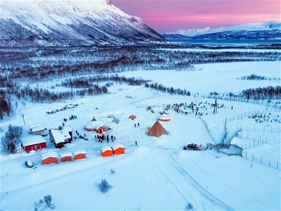 Snødekt landskap med telt, hytter og folk foran fjell.