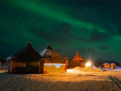 Nordlys over snødekte hytter og lavvo opplyst av varme lys.