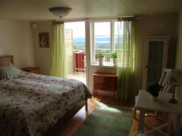 Double bed with floral bedspread in a room with green curtains in front of an open door to the balcony. 