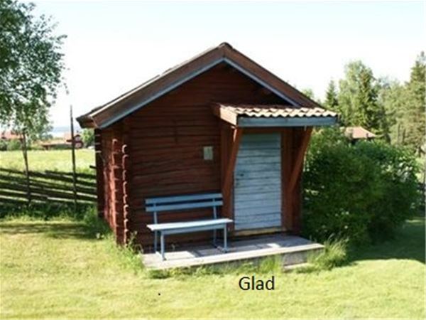 Stuga Glad är en röd liten timmerstuga med blå dörr och en blå bänk bredvid dörren.  