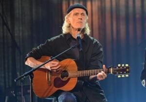 En man sitter på en stol, håller i en gitarr, en miklrofon framför sig, iklädd mörk jacka och skjorta, blå jeans och en svart mössa på huvudet.