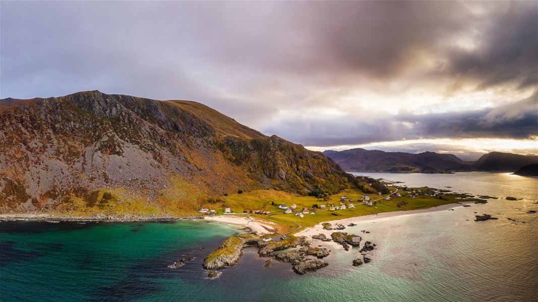 Grotlesanden, Panorama av bratt fjell, lita bygd med hus og grøne jorder ved sandstrand og roleg fjord under skyer..