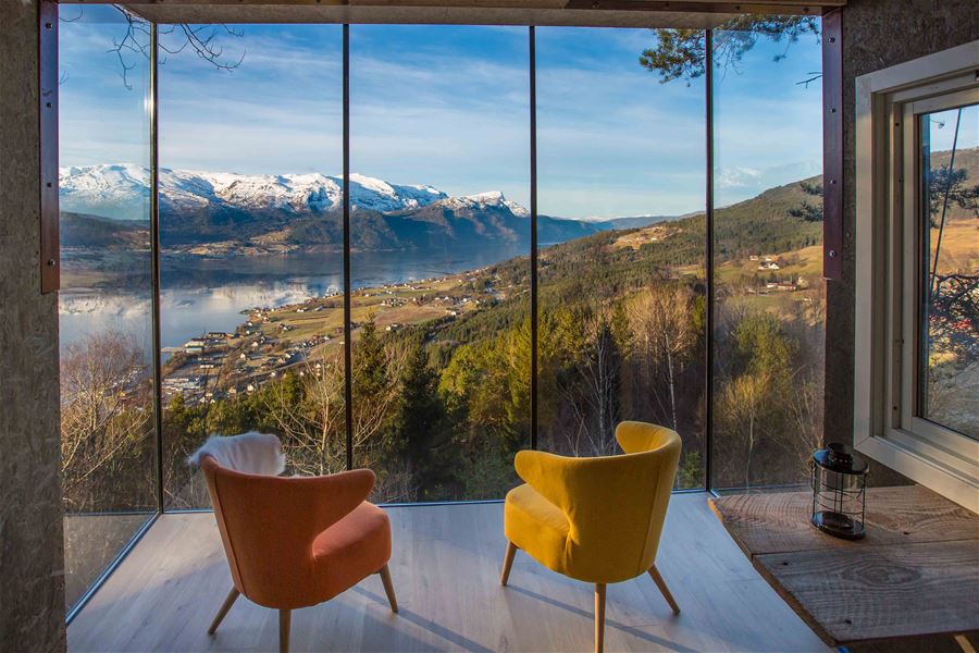 To lenestolar ved store vindauge, med utsikt over fjord, dal, små bygder og snødekte fjell i solskin..