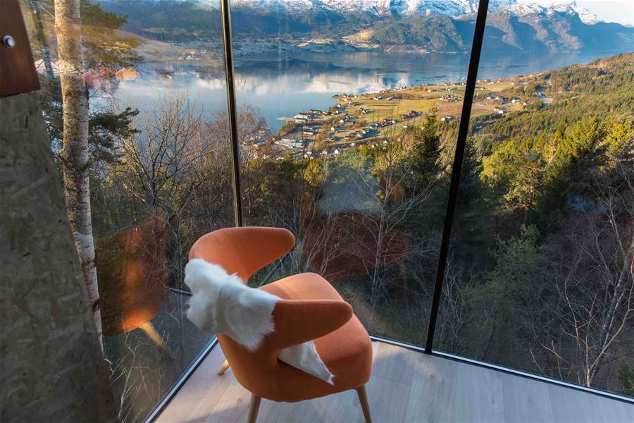 Oransje lenestol med kvitt skinnfell ved stort vindauge, med utsikt over fjord, gardar og snødekte fjell..