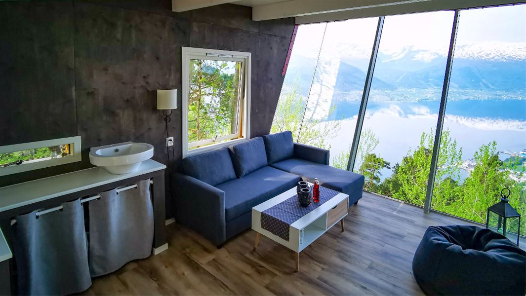 Moderne stoverom med sofa og salbord ved store glasvindauge, med utsyn til fjord, fjell og skog..