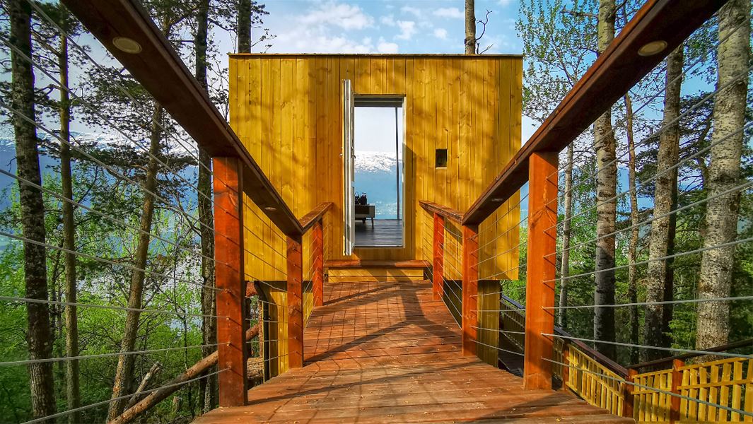 Trebrygge med rekkverk leier til ei gul trehytte i skogen, med fjord og snøkledde fjell synlege i bakgrunnen..