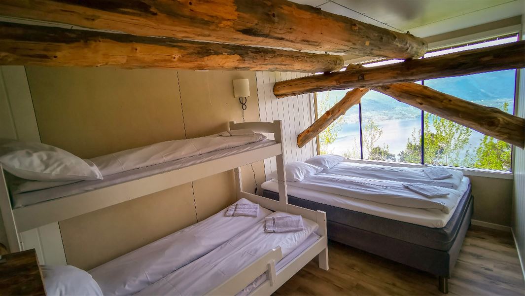 Lite soverom med køyeseng og dobbeltseng, synlege takbjelkar av tre og stort vindauge mot fjord og fjell..