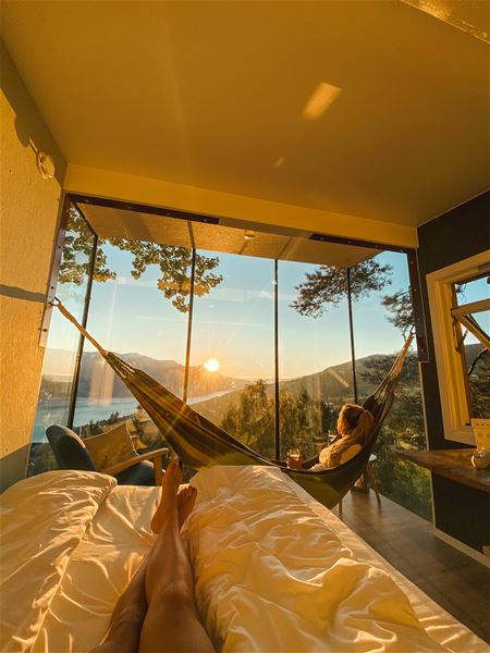 Utsikt frå ei seng mot store glasruter med solnedgang, fjord og fjell, og ei kvinne som kviler i hengekøye..