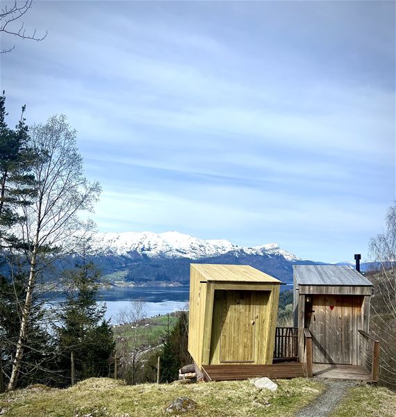To små trehytter på ei grasskledt høgd med grussti, med fjord og snødekte fjell i bakgrunnen..
