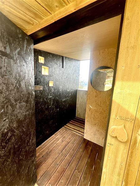 Innsyn i eit lite treinteriør med mørke veggar, små vindauge, rund spegel på vegg og tregolv..