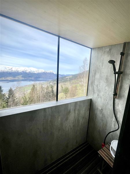 Betongdusj med handdusj og benk, stort vindauge mot fjord, skog og snødekte fjell under blå himmel..
