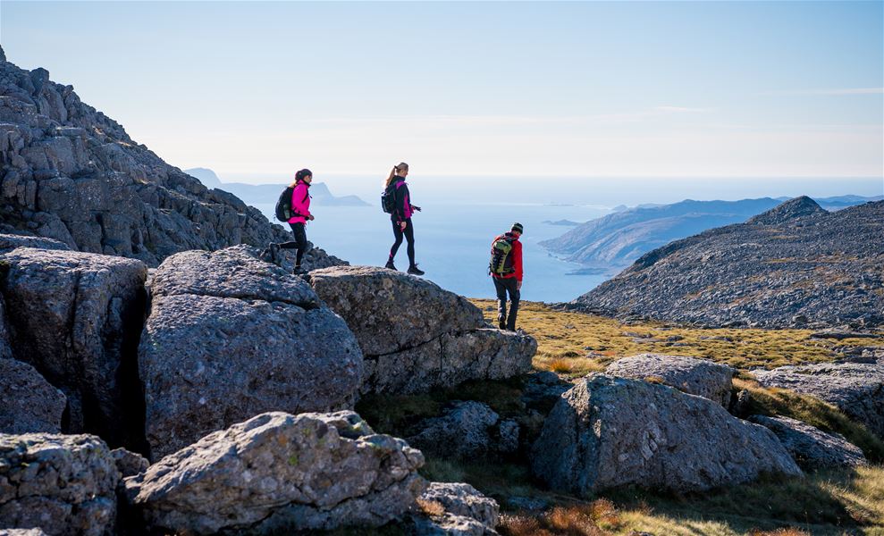 Three hikers with backpacks walk across rocky mountain terrain, overlooking a blue sea and distant islands..