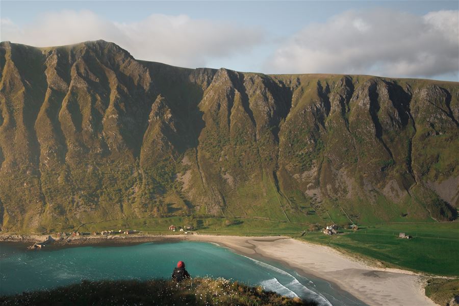 Ein person sit på ein kolle og ser ned mot turkis hav, sandstrand, grøne marker og bratte fjell under skyer..