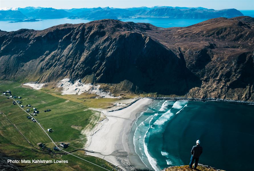 Ein person står på ein fjelltopp og ser ned på ei kvit sandstrand, bølgjer, grøne jorder og bratte fjell ved fjorden..