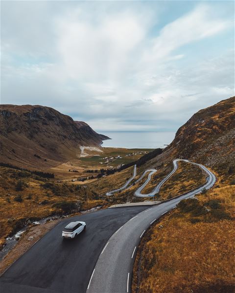 Svingete veg ned ein fjelldal mot fjord og hav, med ein bil i forgrunnen og skyer over fjella..