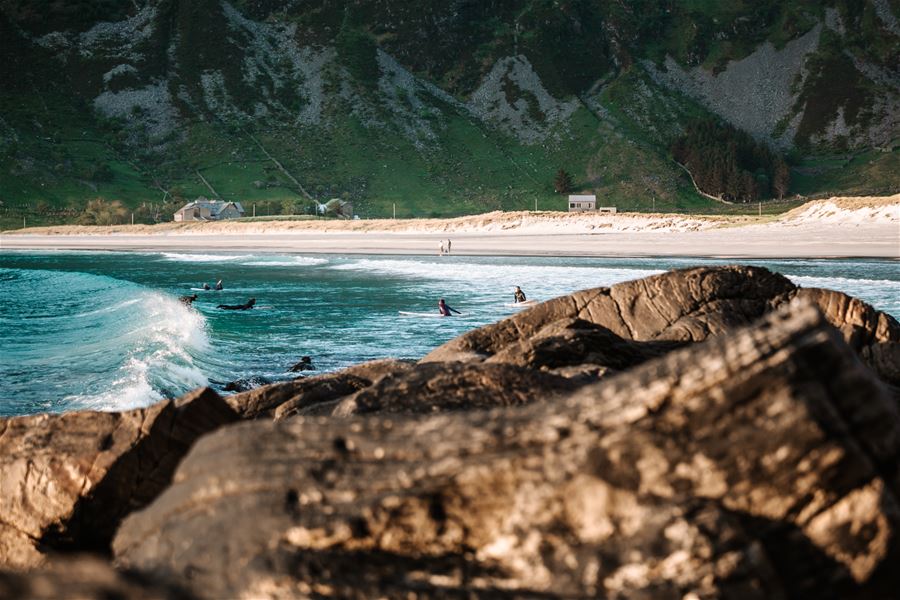 Surfaranar i bølgjene nær store steinar, med sandstrand, små hus og grøne fjell i bakgrunnen..
