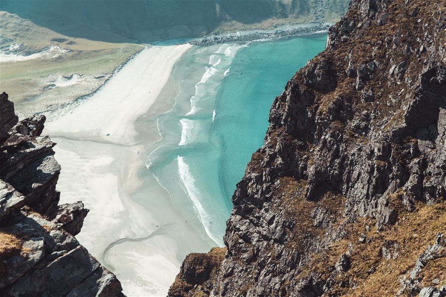 Utsyn frå fjellside ned mot ei lys sandstrand og turkis sjø med bølgjer, omgitt av bratte, steinete klipper..