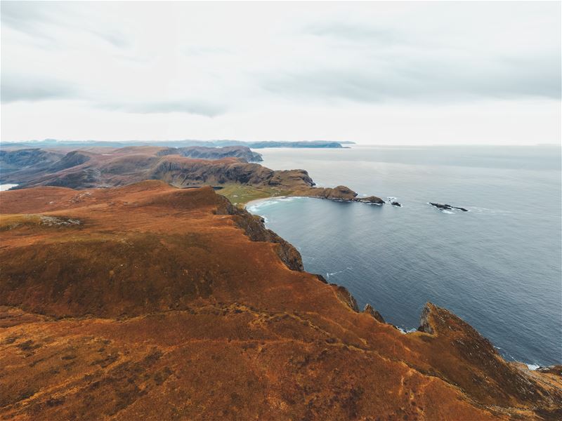 Wide view of rugged brown hills and rocky cliffs above a calm bay and open ocean under an overcast sky..