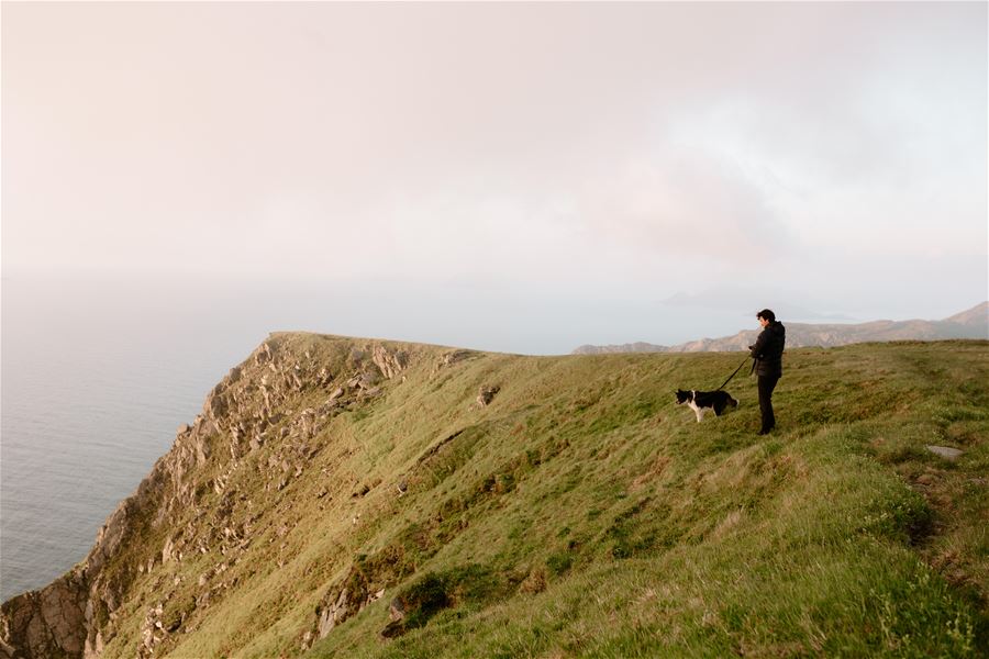 Person stands with a dog on a grassy coastal cliff, overlooking the calm ocean under a hazy sky..