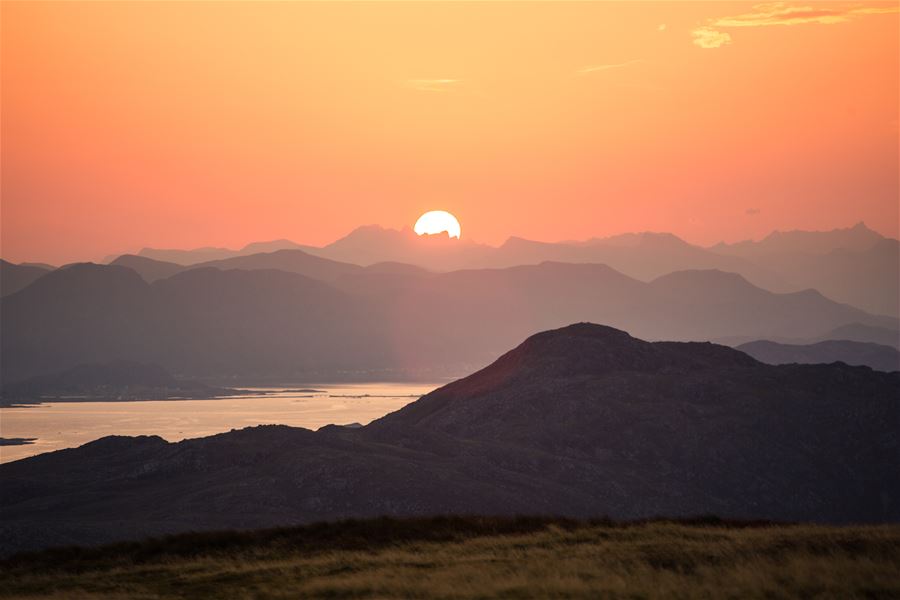 Orange sunset with the sun near the horizon above layered mountain ridges and a distant reflective lake..
