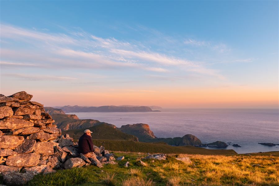 A person sits on rocky ground near a stone pile, overlooking grassy hills and a calm sea at sunset..