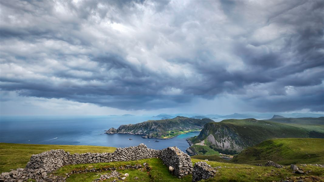 Wide view of rugged green cliffs and a bay below, with stone ruins in the foreground under dark storm clouds..
