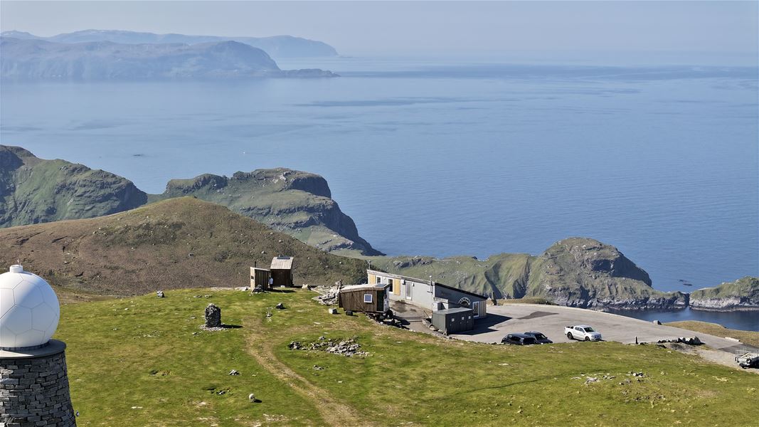 Grassy hilltop with small buildings, a dome structure, and parked cars overlooking rugged green cliffs and calm sea..
