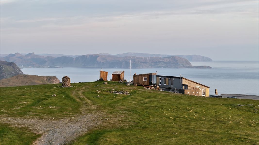 Small cabins and a lodge on a grassy hilltop above calm sea, with distant mountains under a soft evening sky..